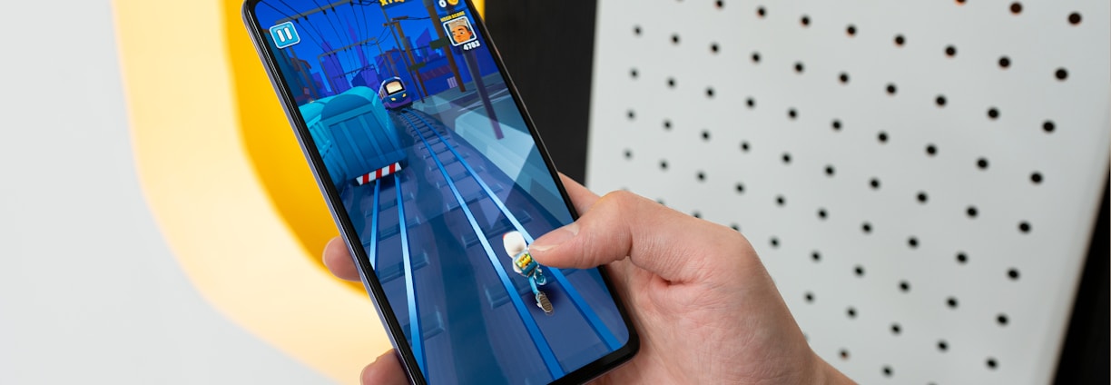 A person is holding a smartphone displaying a mobile game with vibrant graphics. The game depicts a character running on train tracks. The background includes a well-lit wall with a pattern of holes.